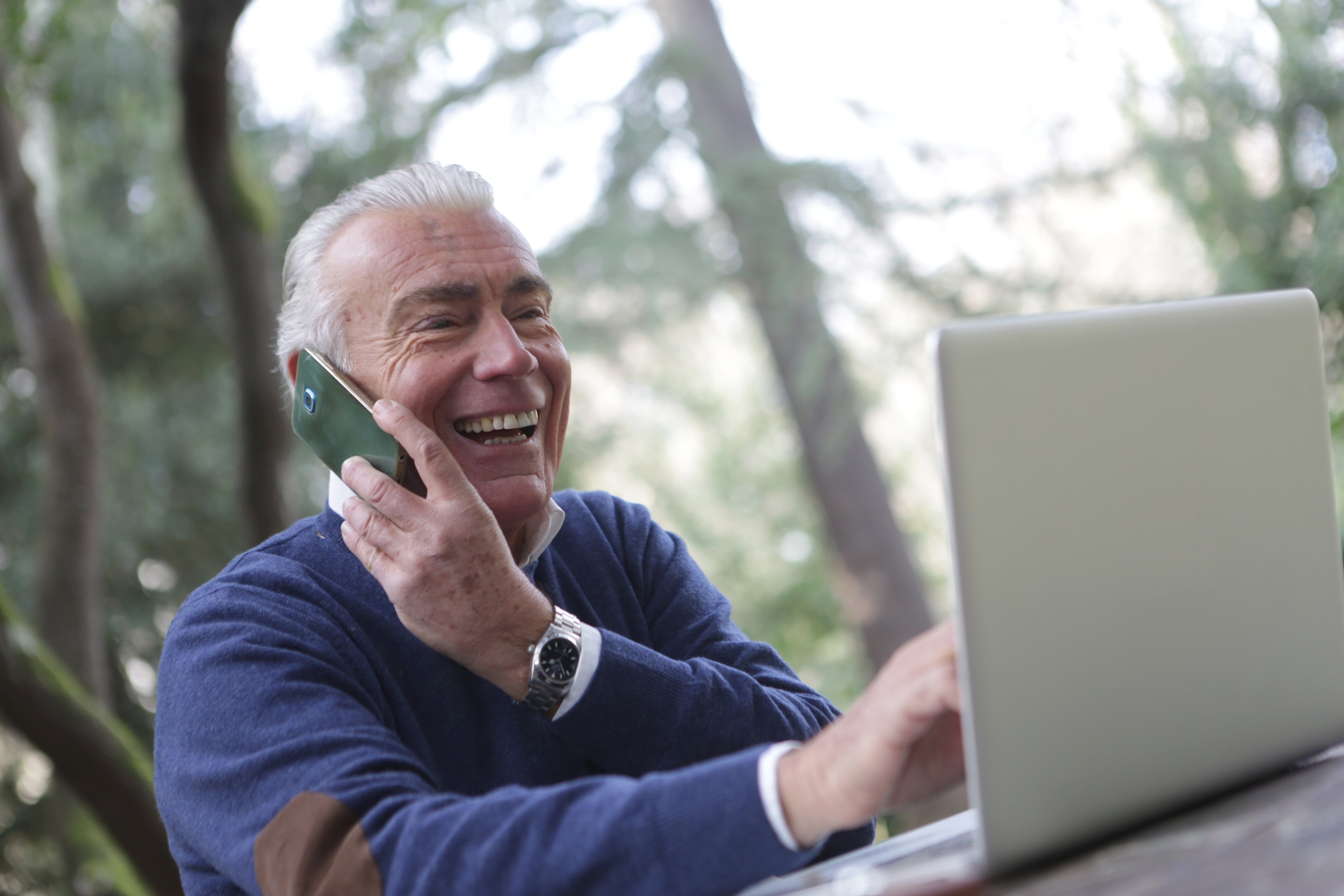 An older man uses a laptop with one hand, while his other holds a smartphone to his ear