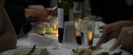 A bottle of wine and plates of food sit on a table as people eat and mingle around it.