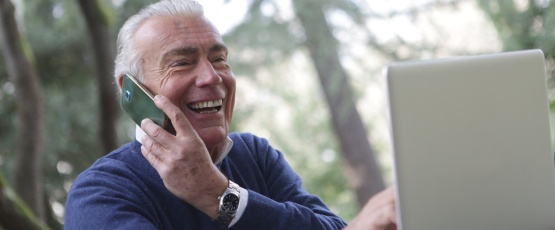 An older man uses a laptop with one hand, while his other holds a smartphone to his ear