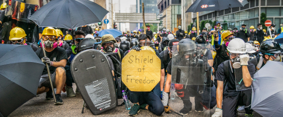 Protesters in Hong Kong crouch in a line in the middle of the street, as if preparing for battle. Most protesters are wearing helmets and gas masks. Many of them are holding makeshift shields and umbrellas. A sign in the center reads "Shield of Democracy"