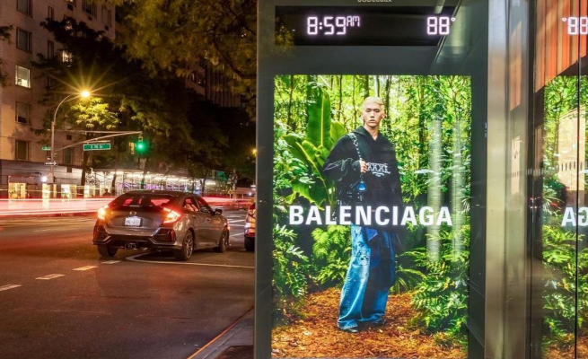 A Balenciaga ad is displayed in a bus stop in New York City, featuring a man in a black windbreaker hoodie against a forest background. 