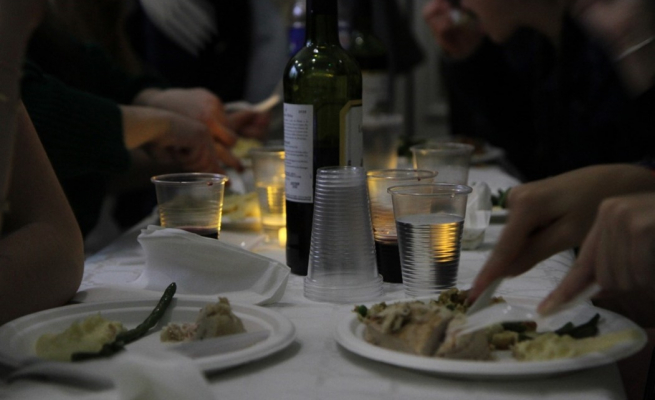 A bottle of wine and plates of food sit on a table as people eat and mingle around it.