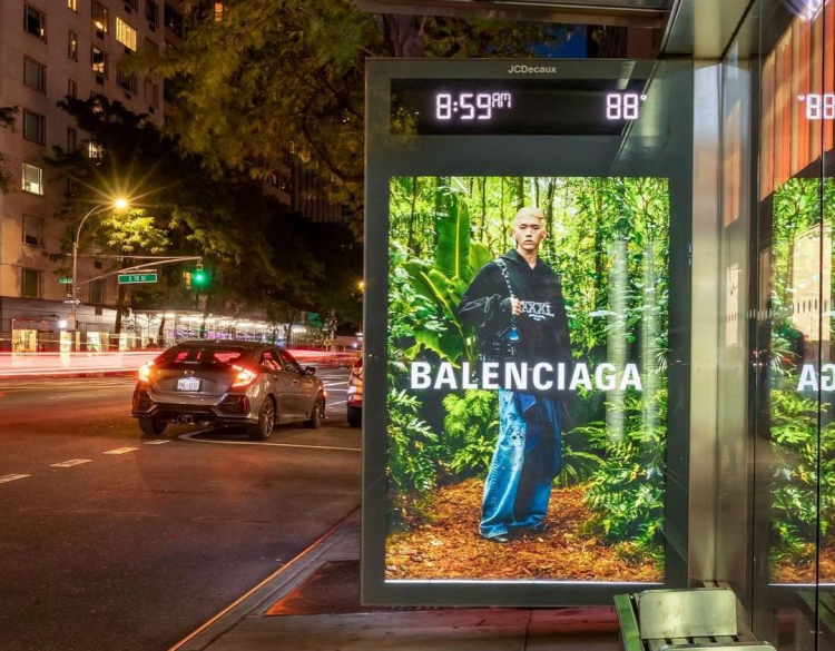A Balenciaga ad is displayed in a bus stop in New York City, featuring a man in a black windbreaker hoodie against a forest background. 