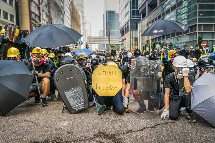 Protesters in Hong Kong crouch in a line in the middle of the street, as if preparing for battle. Most protesters are wearing helmets and gas masks. Many of them are holding makeshift shields and umbrellas. A sign in the center reads "Shield of Democracy"